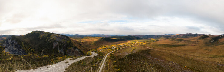 Naklejka premium Panoramic View of Scenic Winding Road surrounded by River, Mountains and Valley in Autumn on a Cloudy Day. Aerial Drone Shot. Taken near Dempster Highway, Yukon, Canada.
