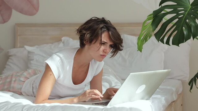 Beautiful smart young freelancer woman wear white t-shirt pajamas lying in bed spending time in bedroom work from home using laptop pc computer in quarantine. Rest relax good mood lifestyle concept