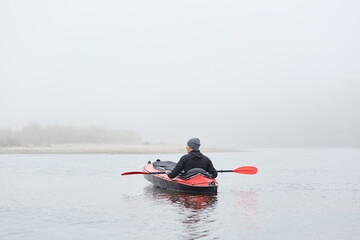 Fototapeta na wymiar Sportsman posing backwards outdoors while sitting in canoe in middle in rover or lake, male looking at foggy nature, enjoying his hobby, doing extreme water sport.