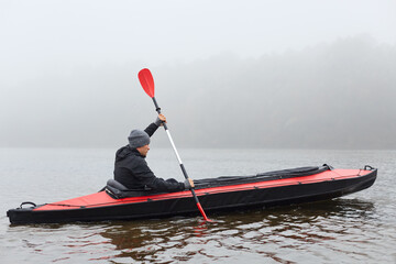 Profile of man floating on lake in kayak, looking concentrated on his way, rowing boat in cold cloudy day, water sport, guy in black jacket padding, pastime in active way. © sementsova321