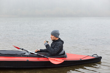 Man paddles red and black kayak and sitting with paddle, holds mug with hot coffee or tea on river...