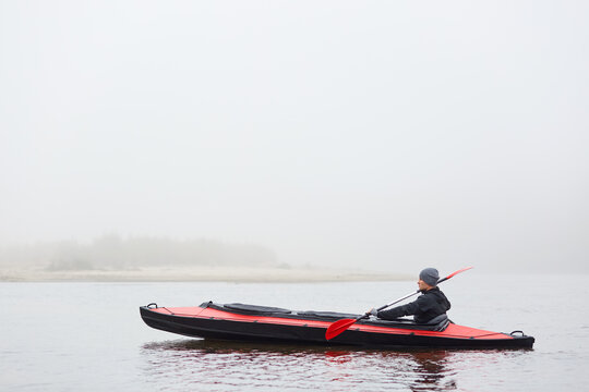 Handsome man paddling canoe on cloudy day, looks concentrated, wearing black jacket and gray cap, holding paddle in hands, spends free time doing water sport and enjoying nature.