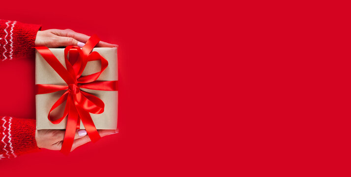 Female hands holding gift box with red ribbon on red background. Top view. Close-up. Banner. Christmas, New Year, Valentine's day and birthday concept.