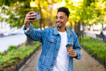 Handsome Afro American man is doing selfie using a smart phone, holding a cup of coffee and smiling while walking in city