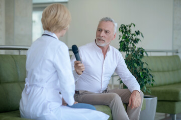 Gray-haired male journalist holding microphone, sitting on sofa, interviewing blonde female doctor
