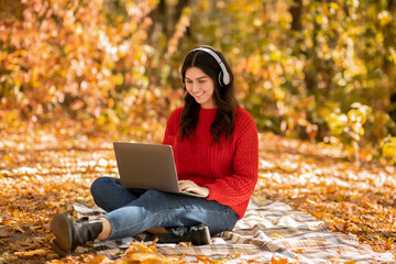 Full length portrait of female student with laptop and headphones participating in online lesson at autumn park