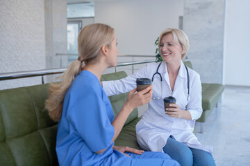Two smiling female medical workers sitting on sofa, drinking coffee