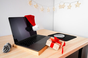 PC on wooden table at home with Santa Claus hat, gift box and xmas decor. Online Christmas shopping concept. Stay at home. Winter holiday preparations