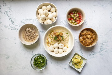 Rice stick noodles with fish ball in white bowl. Garlic cracklings with lard, mined pork, pickled chili, lime and coriander with spring onion various ingredients and condiment on table.