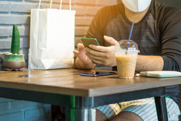 Asian male customer wear face mask using smartphone while sit in waiting social distance queue for take away drink in coffee shop. New normal lifestyle concept. Selective focus on smartphone