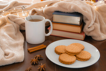 Living room details in winter. Close-up of a white mug with an infusion on a wooden table with anise and cinnamon, a cream colored blanket, cookies and books. Homey winter concept.