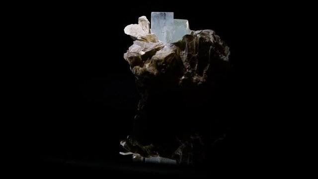 An aquamarine crystal emerging from the matrix of muscovite turns slowly with a reflection of the sample apparent in the glass base. Sample from Argentina.