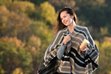 Fototapeta na wymiar Beautiful young woman in a poncho with coffee in hand on nature background. Brunette girl laughing