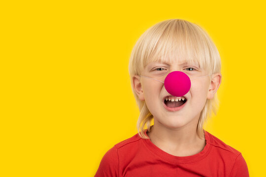 Boy with clown nose discontentedly looking at camera and making faces. Portrait close up on yellow background