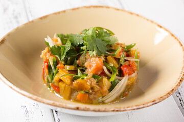 Salad dudlyash. Grilled eggplant, sweet pepper, tomatoes with onion, garlic and fresh herbs in a beige plate on a white background. Restaurant menu. Background image, copy space