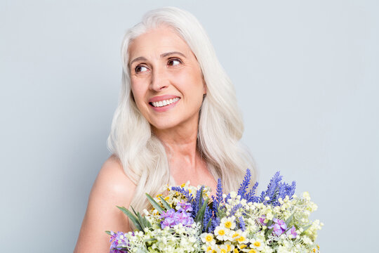 Photo of cute retired woman beaming smile hold big bunch wild flowers look empty space shy isolated grey color background
