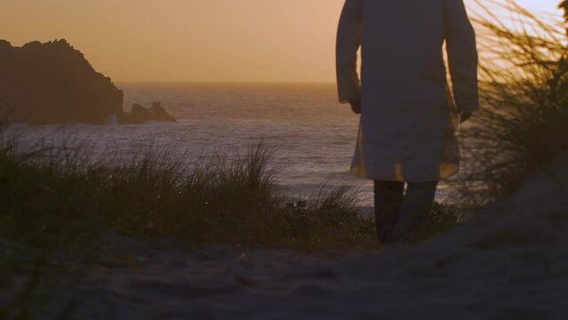Man in kaftan walks beach trail to watch golden sunset, slow motion