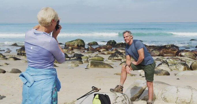 Senior hiker woman taking pictures of senior hiker man using digital camera on the beach.