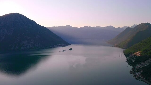 The sun slowly rising from behind a high mountain above the still waters of Kotor Bay, Montenegro, surrounded by hills and mountains,light morning haze above the whole area,cloudless sky, aerial shot.
