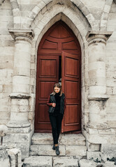 Fototapeta na wymiar young woman in a leather jacket near the entrance to the ancient wooden door of a gothic temple