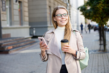 Cheerful young woman wearing coat walking with coffee cup and cellphone