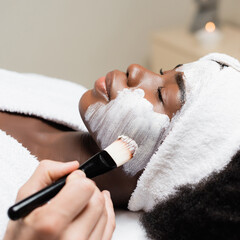 Close up view of spa therapist applying face mask with cosmetic brush on cheek of African American...