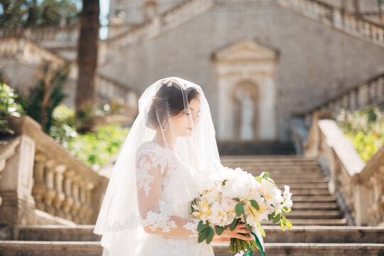beautiful bride covered with a veil with bridal bouquet on the stairs of the Nativity of the Blessed Virgin Mary church in Prcanj