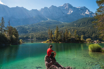 Boy in red shirt sitting on a rock watching a beautiful view over lake Eibsee, in the Bavarian Alps. Famous touristic destination in the middle of the nature in Europe.