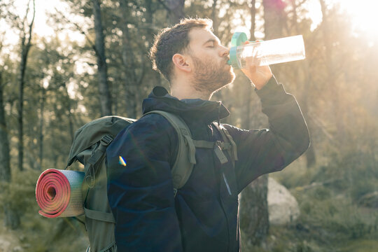 Close-up of young male hiker drinking water from his transparent bottle with background sunlight blurring the image in a mountain forest with soft dark colors.