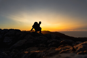 Hiker at the top of a mountain crouching and observing on the mountain ridge a beautiful golden hour sunset with intense color and blue sky fused with yellow, man in profile looking at the sun
