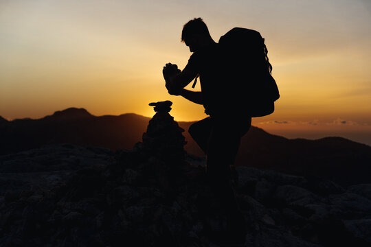 Hiker at the top of a mountain stacking stones in a small hill with a beautiful golden hour sunset illuminating the silhouette, carrying a large backpack