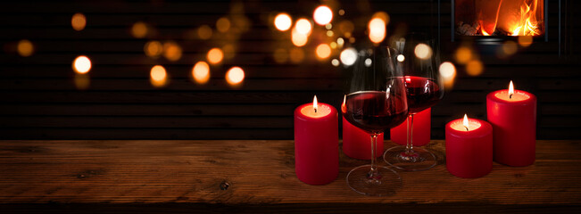 Glasses of red wine with candles on rustic wood in front of a log fire and golden bokeh lights....
