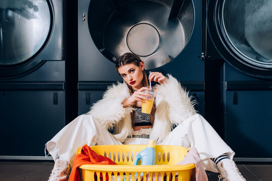 Young Woman Holding Plastic Cup With Orange Juice And Sitting Near Basket With Laundry, Detergent Bottle And Washing Machines In Laundromat