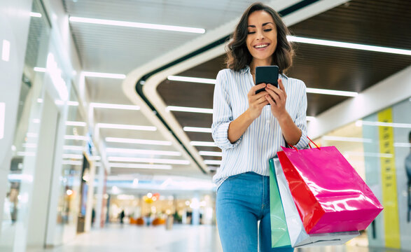 Modern gorgeous young smiling woman in trendy stylish clothes with bright colorful shopping bags is using her smart phone while walking in the mall