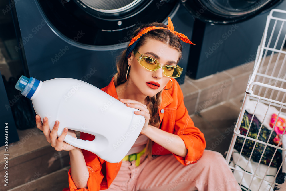 Wall mural high angle view of young woman sitting on floor and holding bottle with detergent in laundromat