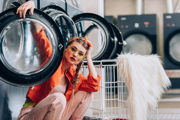 trendy woman in sunglasses sitting near metallic cart and washing machines in laundromat