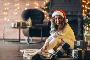 Pretty woman in warm sweater, socks and christmas hat, sitting on the floor at home with gift boxes