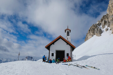 Silhouettes of sitting skiers next to chapel at feet of huge rocks in mountains on sunny day. Around refuge Locatelli around Tre Cime Peaks. Dolomites, Italy