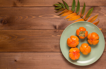 Abstract autumn still life: yellowed leaf and ripe persimmon in plate on a brown wooden table. Flat lay. Copy space