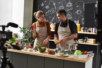 Happy young couple in aprons standing in the kitchen and discussing the new recipe together they...