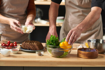 Close-up of couple in aprons taking ingredients and preparing food together at the table