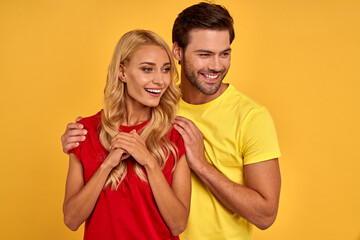 Smiling young couple friends guy girl in red yellow t-shirts posing isolated on yellow wall background studio portrait. People emotions lifestyle concept. Mock up copy space. Hugging, looking aside.