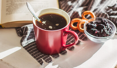 A Cup of hot steaming coffee in a red mug, cookies, and an old book on a white wooden table