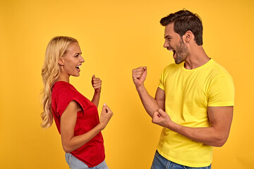Happy young couple two friends guy girl in red yellow t-shirts isolated on yellow background studio. People lifestyle concept. Mock up copy space. Clenching fists like winner, looking at each other.