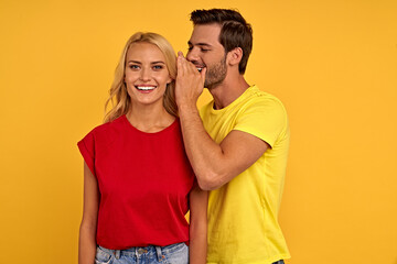 Fototapeta premium Excited young couple friends guy girl in casual clothes isolated on yellow background. People lifestyle concept. Mock up copy space. Whispers gossip and tells secret behind hand, doing winner gesture