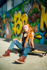 Fashion lifestyle, Beautiful young redhead  woman with skateboard sitting near the wall with graffiti at the day time.