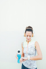 Naklejka premium young female athlete looks at watch while carrying energy drink and listening to music with headphones