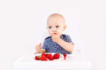 Baby girl sitting in high child's chair eating fruits on a white background. Baby food concept, space for text