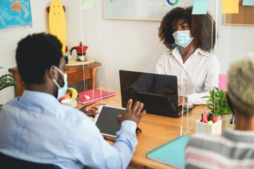 Fototapeta premium African co-workers using laptops inside creative office behind safety glass wearing protective face masks - Business and social distance concept - Focus on girl face