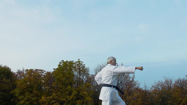 Against the background of the sky and trees, an old man athlete trains formal karate exercises